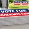 Stack of 24x18 corrugated plastic yard signs on a sign shop table with staked signs on the lawn outside